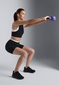 Musta urheilullinen crop-top ja shortsit valkoisilla yksityiskohdilla, jota pitää päällään henkilö, joka tekee kyykkyä purppurakettlebelli kädessä harmaalla taustalla.