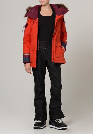Helle orange Parka mit einer mit Fell gefütterten Kapuze, lila Innenfutter, bunten gemusterten Akzenten, kombiniert mit schwarzen wasserdichten Hosen und schwarzen Turnschuhen.
