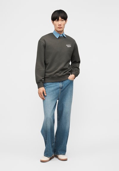 Νεαρός άνδρας που φοράει σκούρο πράσινο φούτερ με το λογότυπο "Maison Kitsuné", ανοιχτό μπλε τζιν παντελόνι με φαρδιά μπάσκα, λευκά παπούτσια, στέκεται με το ένα χέρι στην τσέπη.