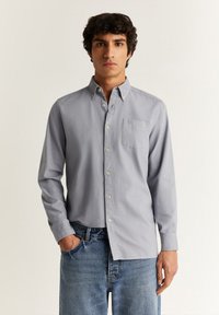Camicia a maniche lunghe grigia chiara con bottoni, realizzata in tessuto morbido, con taschino sul petto e bottoni bianchi; indossata con jeans blu.
