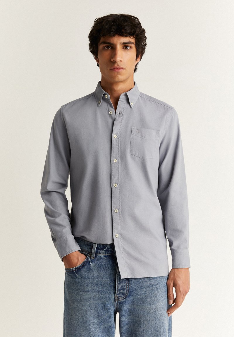 Camicia a maniche lunghe grigia chiara con bottoni, realizzata in tessuto morbido, con taschino sul petto e bottoni bianchi; indossata con jeans blu.