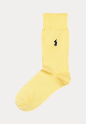 En enkelt gul sok med ribbet kant og broderet navyblåt polospillerlogo på anklen mod hvid baggrund.