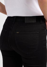 Svarta denimjeans med en slät yta, med en klassisk femfickdesign och en gyllene "Lee"-logotyp på bakfickan. Svart midjebandsetikett.