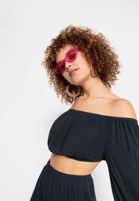 Kattögon-solglasögon i genomskinlig rosa, bärs med en svart off-shoulder crop top. Modellen har lockigt hår och stora guldörhängen.