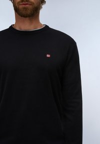 Homme barbu portant un pull noir à col rond avec un petit emblème de drapeau rouge et blanc sur la poitrine gauche, sur fond uni.