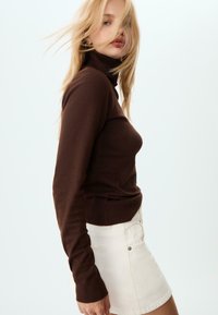 Brauner Rollkragenpullover mit langen Ärmeln, gerippten Bündchen und taillierter Form. Kombiniert mit einem weißen Minirock.