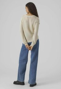 Maglione a maglia beige chiaro con un motivo testurizzato, abbinato a jeans blu larghi e scarpe con tacco nere. Vista posteriore, mani unite.