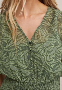 Blusa verde a motivo con scollatura a V, chiusura con bottoni davanti e un tessuto leggero e testurizzato con un design a onde.