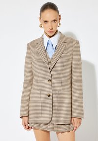 Beige takki ruudullisella kuviolla, jossa on muodollinen istuvuus, kultaiset napit, kaksi etutaskua ja yksityiskohtainen kaulus. Yhdistetty vaaleansiniseen solmioon ja poimutettuun hameeseen.