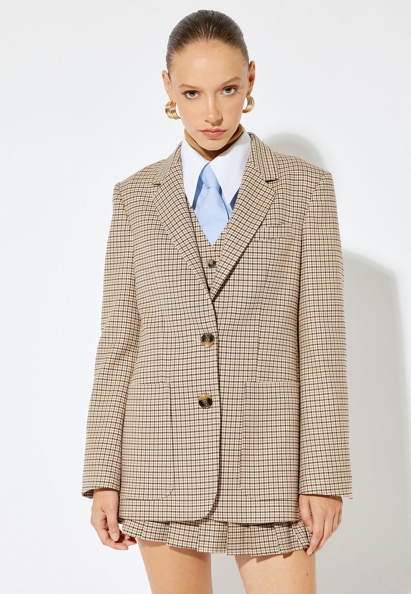 Beige takki ruudullisella kuviolla, jossa on muodollinen istuvuus, kultaiset napit, kaksi etutaskua ja yksityiskohtainen kaulus. Yhdistetty vaaleansiniseen solmioon ja poimutettuun hameeseen.