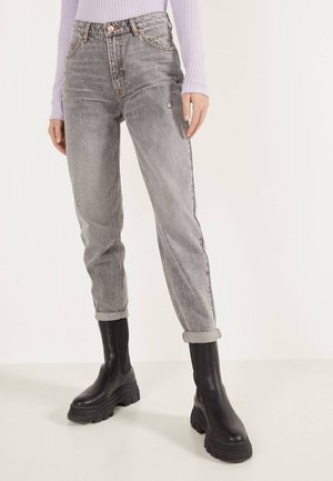 Person iført slidte grå high-waist jeans med opbuk og sorte chunky plateau-støvler, kombineret med en lyse lilla ribstrikket sweater.