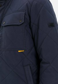 Marineblau gesteppte Jacke mit einer großen Klappentasche mit schwarzem Zubehör, einer Seitentasche und einem auffälligen gelben Etikett auf der Vorderseite.
