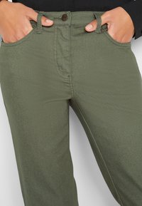 Pantalon vert olive en tissu doux, avec un design classique à cinq poches, fermeture à boutons à l'avant et passants pour ceinture. Texture lisse.