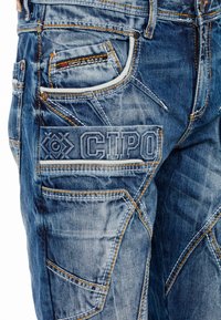 Blauwe denim cargo broek met oranje stiksels, meerdere zakken en reliëflogo-details. Gehavend textuur met een aansluitend ontwerp.