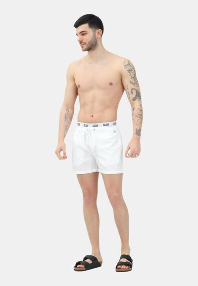 Hvide svømmedragter med snøre og grå elastisk talje. Båret sammen med sorte sandaler, der har en glat overflade og et enkelt design.