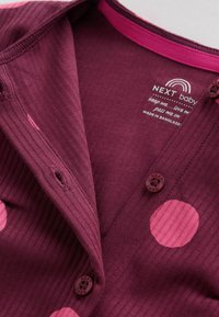 Gros plan sur un cardigan bébé côtelé bordeaux avec de grands pois roses, des boutons et un texte de marque imprimé en blanc à l'intérieur du col.