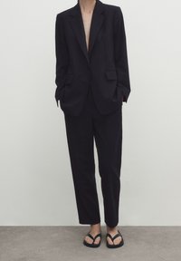 Zwarte op maat gemaakte blazer met scherp uitgesneden revers, gecombineerd met hooggesneden zwarte broek. Draag met zwarte flip-flops. Glad stoftextuur.