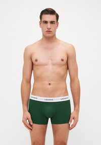 Zeleni pamučni bokserice s bijelim elastičnim pojasom na kojem je istaknut logotip "LACOSTE", uski dizajn bez dodatnih uzoraka ili akcenta.