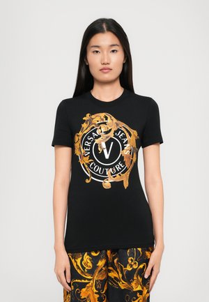 Donna che indossa una t-shirt nera di Versace Jeans Couture con stampa barocca dorata e pantaloni neri abbinati con pattern ornate dorate.