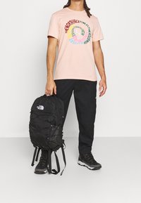 Světlé růžové tričko s barevným kruhovým logem, černý batoh s bungee provazy a logem, černé kalhoty a tmavé turistické boty.