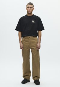 T-shirt noir à manches courtes avec un logo sur la poitrine, associé à un pantalon ample couleur sable et des baskets noires. Le tissu semble lisse et résistant.