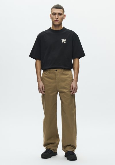 T-shirt noir à manches courtes avec un logo sur la poitrine, associé à un pantalon ample couleur sable et des baskets noires. Le tissu semble lisse et résistant.