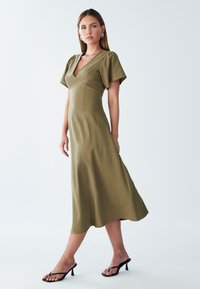 Ženska stoji v olivno zeleni midi obleki s kratkimi rokavi in V-vratom, obuta v črne pete, pred enotno ozadje.