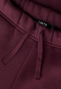 Detail krajkovaného pasu vínových tepláků s odpovídajícími copánkovými šňůrkami a viditelnou černou štítkem s nápisem "LMTD."