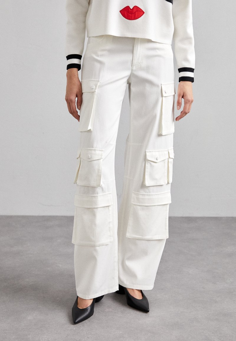 Pantalons cargo blancs à jambes larges avec plusieurs poches, associés à un pull blanc cropped arborant des lèvres rouges, complétés par des escarpins noirs à bout pointu.