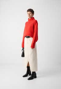 Roter gestrickter Rollkragenpullover mit Rippdetails, kombiniert mit einem hellen Wickelrock und schwarzen, quadratisch geschnittenen Stiefeletten. Kleine schwarze Handtasche.