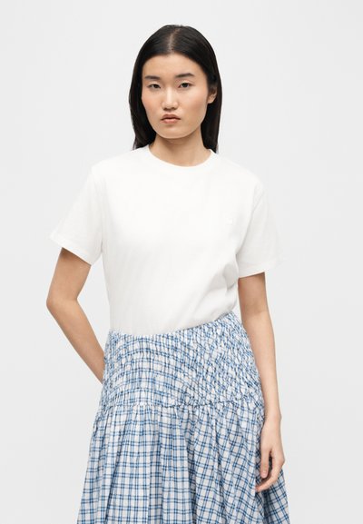 Femme aux longs cheveux bruns portant un haut blanc à manches courtes uni et une jupe taille haute à carreaux bleus et blancs, debout devant un fond blanc.