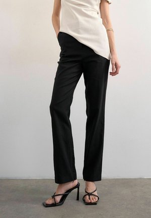 Femme portant un pantalon noir droit, un top asymétrique beige, des sandales noires à talons hauts et brides, et un bracelet doré au bras, devant un mur uni.