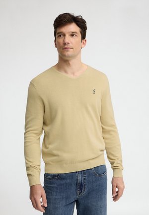 Uomo che indossa un maglione beige a V con un piccolo logo ricamato di un golfista sul petto e jeans blu, su sfondo chiaro e semplice.