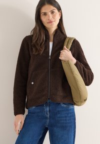 Femme portant une veste polaire zippée marron foncé, un jean bleu, et tenant un sac à bandoulière matelassé beige devant un mur clair uni.