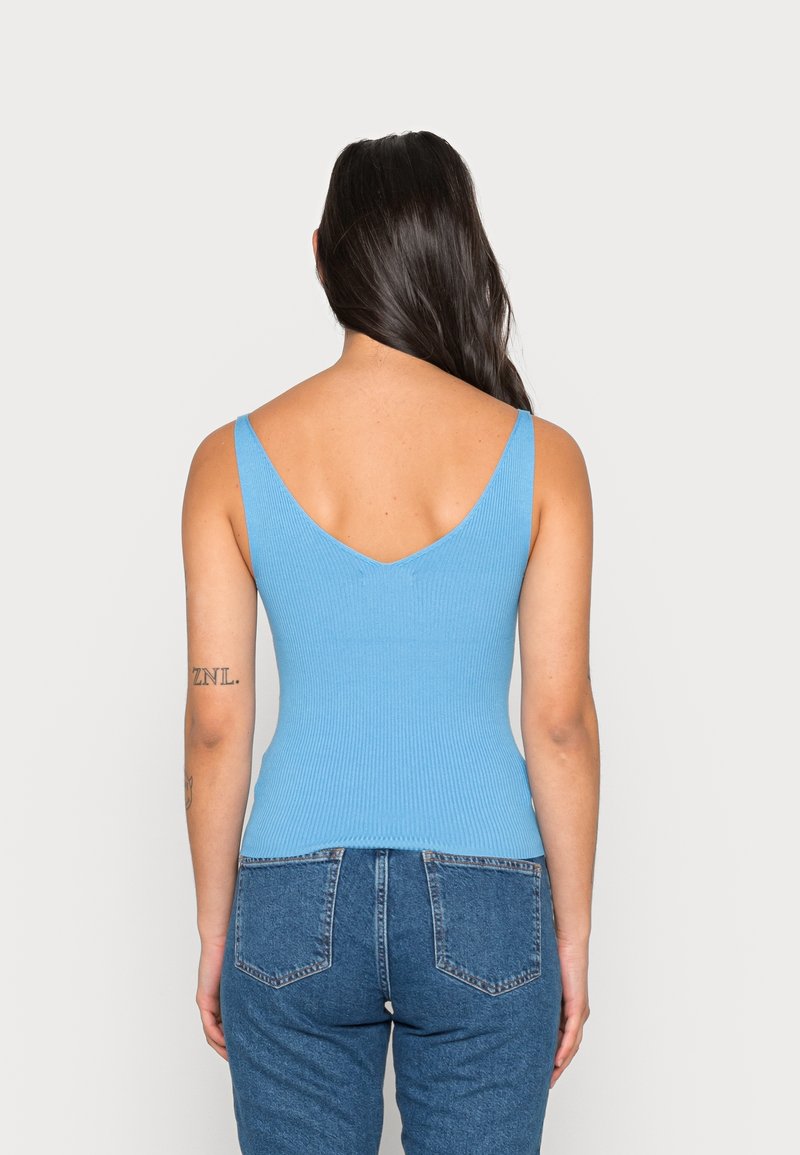 Femme aux longs cheveux foncés portant un top côtelé bleu clair sans manches et un jean bleu, vue de dos sur un fond uni.
