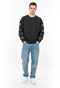 Felpa grigia scura con varie grafiche colorate sulle maniche, abbinata a jeans larghi blu chiaro e sneaker bianche. Tessuto morbido nel complesso.