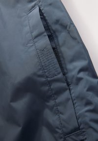 Nepremokavá námornícka modrá bunda s hladkou textúrou, vybavená bočným vreckom so zipsom a prešitými detailmi. Ľahký dizajn.