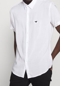 Man som bär en vit kortärmad skjorta med knappar och en liten svart logotyp på vänster bröst, matchat med svarta byxor.