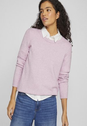 Pull rose clair à manches longues, décolleté rond et poignets côtelés, porté par-dessus une chemise blanche à col. Tissu tricoté doux et texturé.