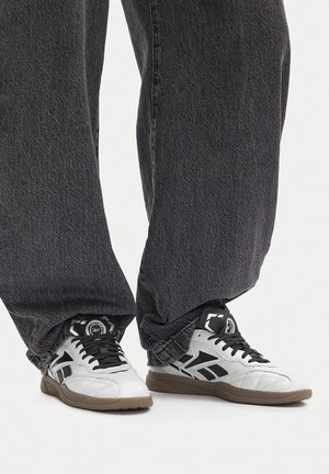 Sneakers bianche e nere con suole in gomma indossate sotto jeans neri lavati e ampi, su uno sfondo neutro.