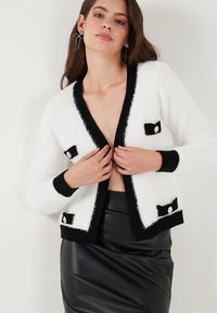 Valkoinen ja musta neulecardigan, jossa on pörröinen pinta, mustat yksityiskohdat, nauhataskujen koristeena ja suuret koristepainikkeet.