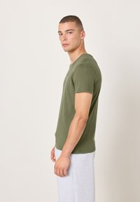 Alyvuogių žalia, trumpomis rankovėmis marškinėliai su apvalia iškirpte, pagaminti iš minkštos medžiagos. Dėvimi kartu su pilkais sportiniais kelnytemis, rodant šoninį profilį.