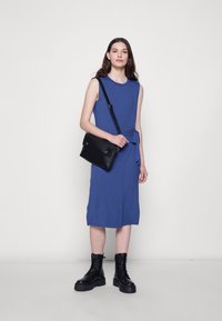 Vestido azul sem mangas, até ao joelho, em tecido suave, com um detalhe de nó lateral. Combinado com uma mala preta de ombro e botas de plataforma com atacadores.