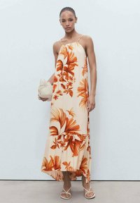 Robe maxi à motifs floraux en orange et beige, avec des bretelles spaghetti et un ourlet volanté. Le modèle tient un sac en osier et porte des sandales blanches.