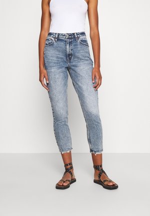 Femme portant un jean court taille haute bleu clair avec des ourlets effilochés, un top blanc sans manches et des sandales plates noires à brides cloutées.