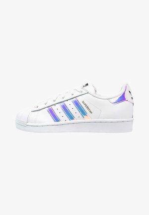 Zapatillas adidas Superstar blancas con rayas iridiscentes azules, un capuchón de goma texturizado y una suela plana blanca. Diseño clásico con cordones.