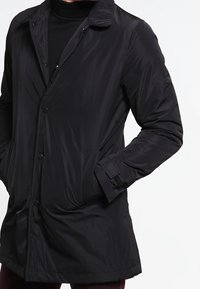 Černý péřový kabát s knoflíkovým zapínáním, kulatým límečkem, bočními kapsami a texturovaným povrchem. Má volný střih a dlouhé rukávy.