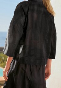 Kvinde iført løs sort jakke og shorts med subtile lodrette striber, står nær udendørs pool med havet i baggrunden.