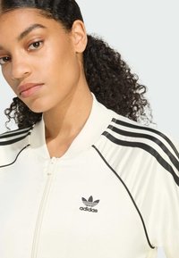 Chaqueta blanca con cremallera y logo negro de Adidas, y tres rayas negras en cada hombro. Tejido suave, cuello estilo bomber.