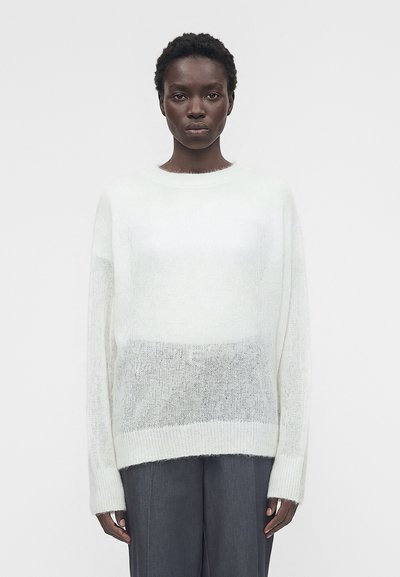 Personne aux cheveux courts et bouclés portant un pull en maille blanc ample et un pantalon gris foncé, debout devant un fond uni.